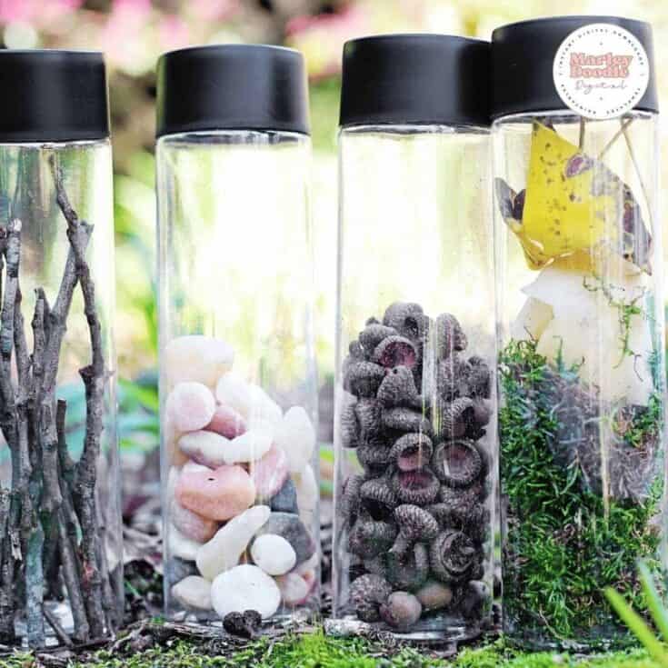 Four clear plastic sensory bottles with black lids lined up outside on soil and moss. The bottles are filled with nature finds like small sticks, smooth pink and white pebbles, dark seed pods, and green moss with a yellow leaf.