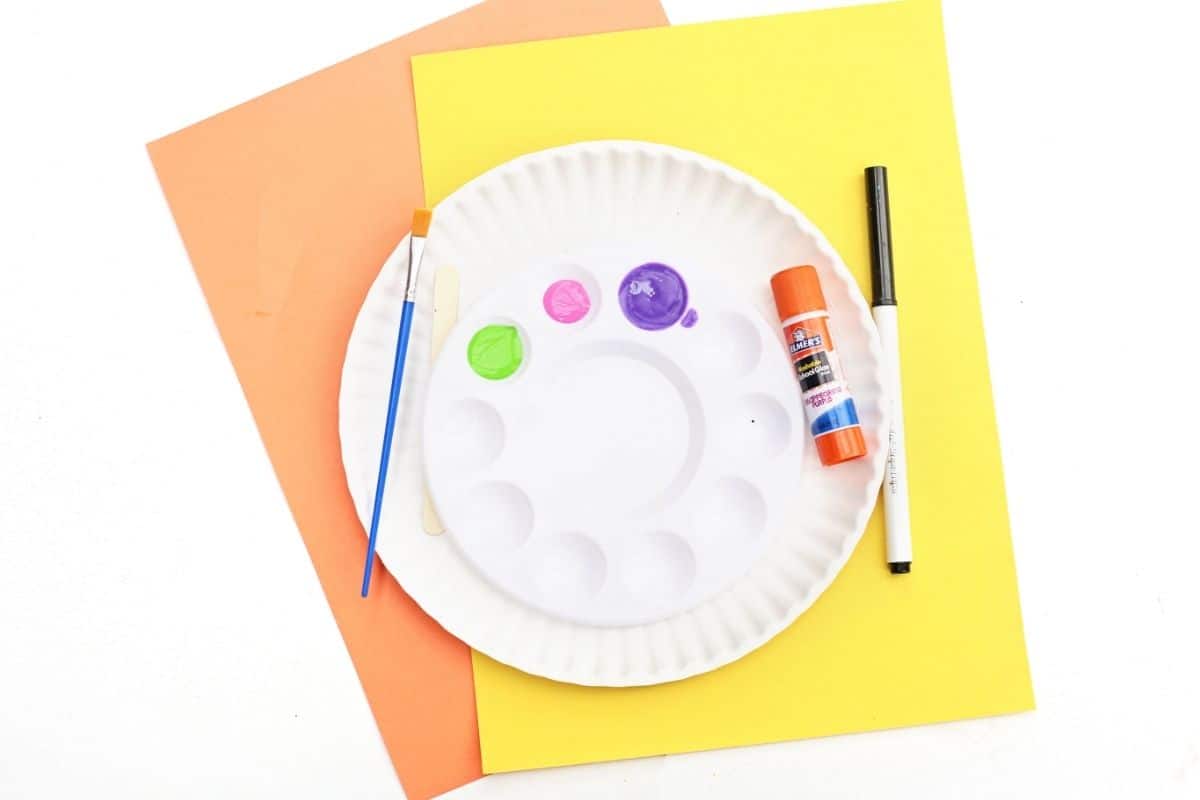 Overhead image of kids’ craft supplies arranged on orange and yellow construction paper, including a white paint palette with green, pink, and purple paint, a flat paintbrush, popsicle stick, glue stick, black marker, and a white paper plate. The setup suggests preparation for a colorful DIY or art project.