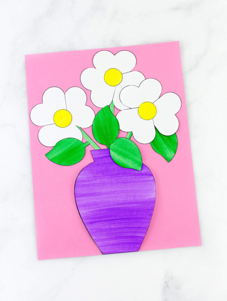 Paper craft of a purple vase holding five white flowers with yellow centers and green leaves, all glued onto a pink background. The vase and flowers appear hand-colored, suggesting a kid-friendly art project.