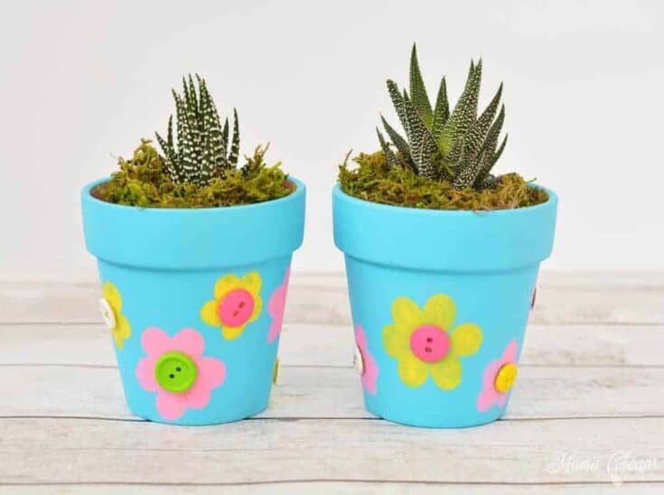 Two small succulents planted in bright blue flower pots decorated with colorful foam flowers and button centers. The pots sit on a light wooden surface and are topped with moss, creating a cheerful, kid-friendly DIY plant gift.