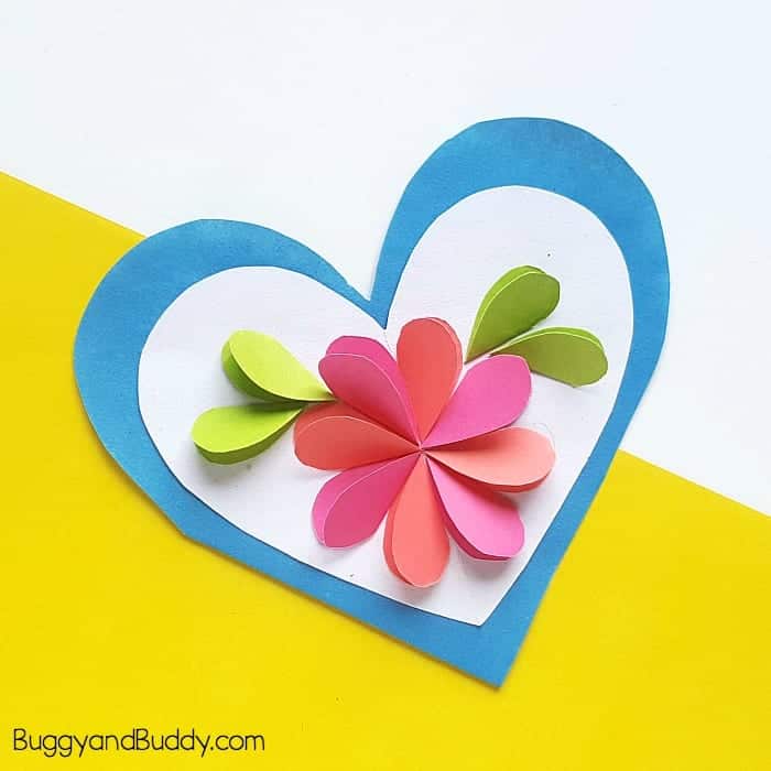 A handmade heart-shaped card features a layered blue and white heart background with a 3D paper flower in the center. The flower has pink and coral petals with green leaves, all folded for a dimensional effect. The card is displayed on a white and yellow diagonal background and labeled with “BuggyandBuddy.com” in the corner.