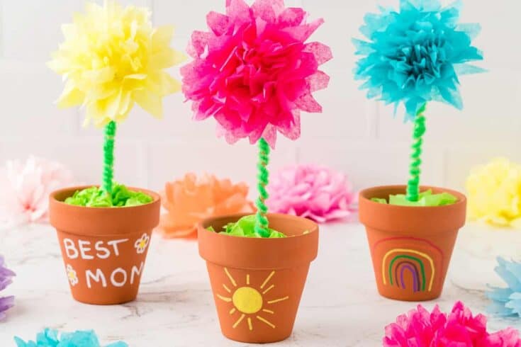 Three colorful tissue paper flowers with green pipe cleaner stems displayed in small terracotta pots decorated with hand-drawn designs like “Best Mom,” a sun, and a rainbow. Each flower is a different color—yellow, pink, and blue—making it a bright and cheerful craft perfect for gifting.