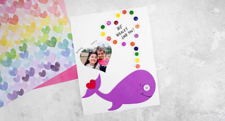 A handmade card features a purple paper whale with a white button eye and a red heart near its tail, blowing a stream of colorful buttons upward. Above the whale, handwritten text reads “We whaley love you!” with a heart-shaped photo of two smiling children labeled Annabelle and Maddie. The card is set on a white background with pastel construction paper peeking from the side.
