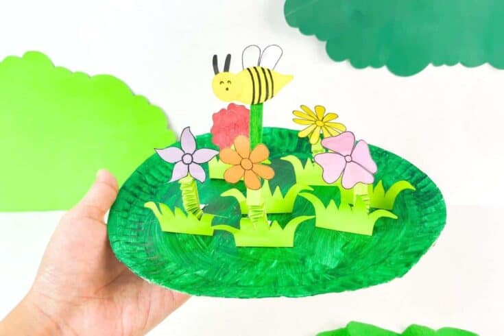 Hand holding a paper plate garden craft painted green, featuring pop-up paper flowers, grass, and a smiling yellow bee with black stripes. The 3D elements are made from folded paper and pipe cleaners, creating a playful spring or nature scene.