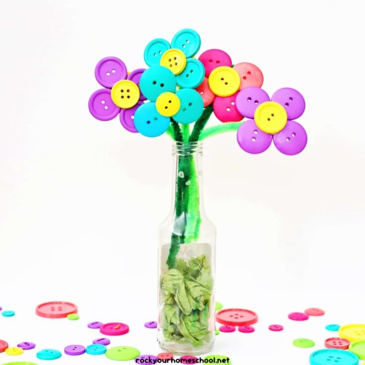 Colorful button flowers with green pipe cleaner stems arranged in a clear glass bottle, with green tissue paper inside to resemble leaves. Each flower is made from brightly colored buttons forming petals and centers, creating a cheerful handmade bouquet.
