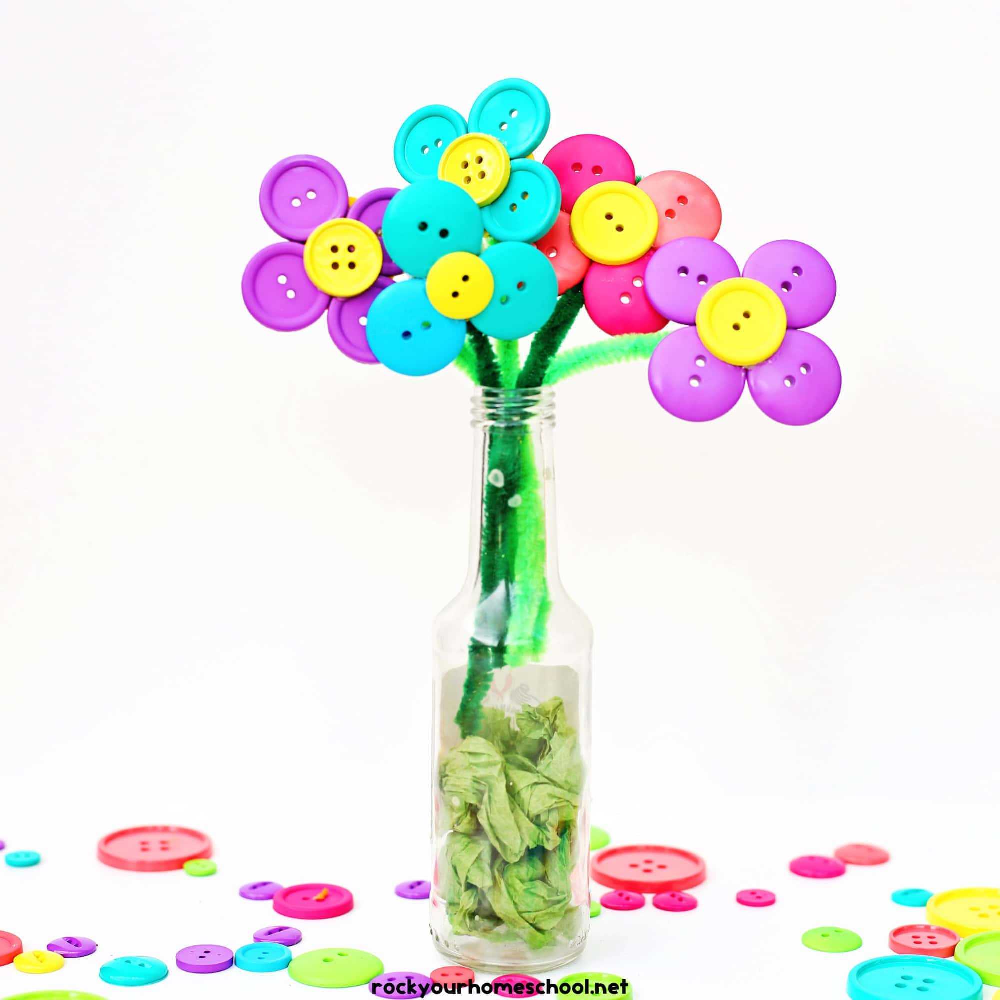 Colorful button flowers with green pipe cleaner stems arranged in a clear glass bottle, with green tissue paper inside to resemble leaves. Each flower is made from brightly colored buttons forming petals and centers, creating a cheerful handmade bouquet.