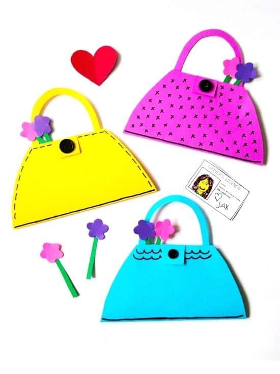 Three colorful paper purse crafts in yellow, pink, and blue are decorated with buttons, hand-drawn details, and foam flowers sticking out of the tops. A small illustrated “driver’s license” labeled with a child’s drawing and the name “Jax” is placed next to the blue purse. The scene is arranged on a white background with a red paper heart and extra foam flowers scattered around.