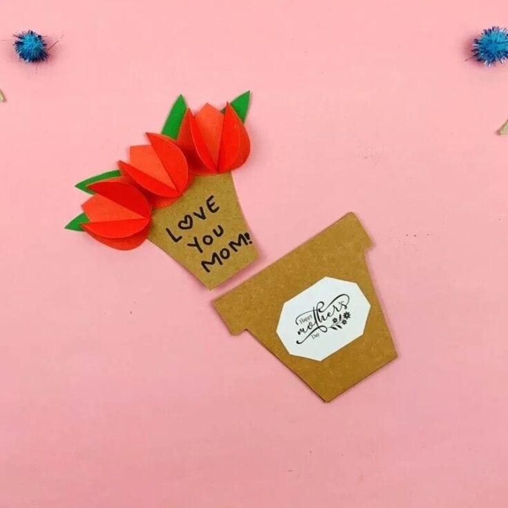 A handmade Mother's Day card shaped like a flower pot opens to reveal three 3D red paper tulips with green leaves and the handwritten message “Love You Mom!” on the inside. The outer flower pot has a white label that reads “Happy Mother’s Day” in script. The card is displayed on a pink background with small blue pom-poms for decoration.