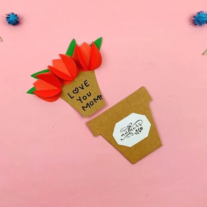 A handmade Mother's Day card shaped like a flower pot opens to reveal three 3D red paper tulips with green leaves and the handwritten message “Love You Mom!” on the inside. The outer flower pot has a white label that reads “Happy Mother’s Day” in script. The card is displayed on a pink background with small blue pom-poms for decoration.