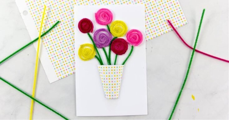 A handmade card features a bouquet of spiral flowers crafted from colorful pipe cleaners in shades of pink, purple, yellow, and red with green stems. The flowers are arranged inside a polka dot paper vase and glued onto a plain white card. The background includes matching polka dot paper and craft supplies.