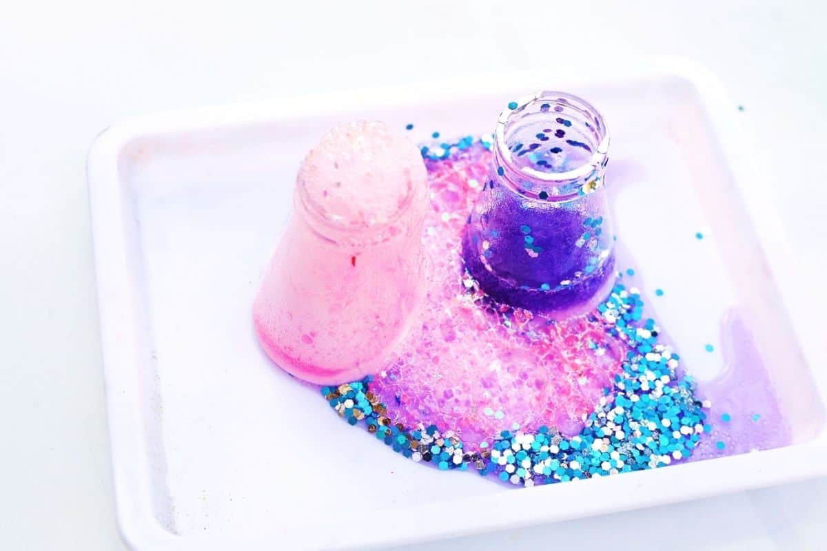 Two small flasks sit in a white tray as bright pink and deep purple foam bubble up and spill over into a sparkling layer of blue and silver glitter. The colorful fizzy reaction spreads across the tray, showcasing the exciting hands on Easter STEM activity in action.