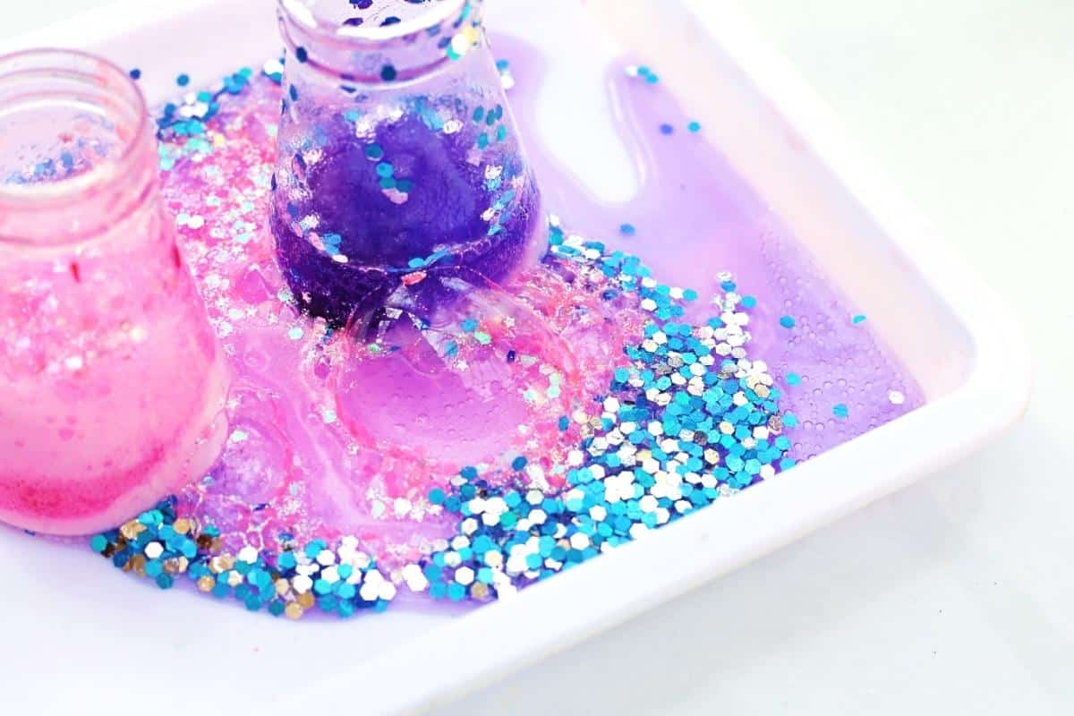 Close up of two small flasks bubbling over with bright pink and deep purple foam inside a white tray, surrounded by chunky blue and silver glitter. The fizzy liquid spills and mixes together, capturing the colorful chemical reaction of the Easter themed STEM activity in action.