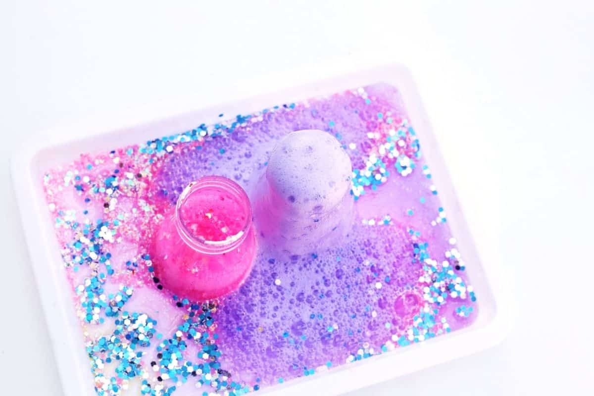 Overhead view of two small flasks in a white tray as bright pink and pale purple foam bubble up and spread across a sparkling layer of blue and silver glitter. The colorful fizz fills the tray with swirling foam, capturing the exciting chemical reaction of the Easter themed STEM activity.