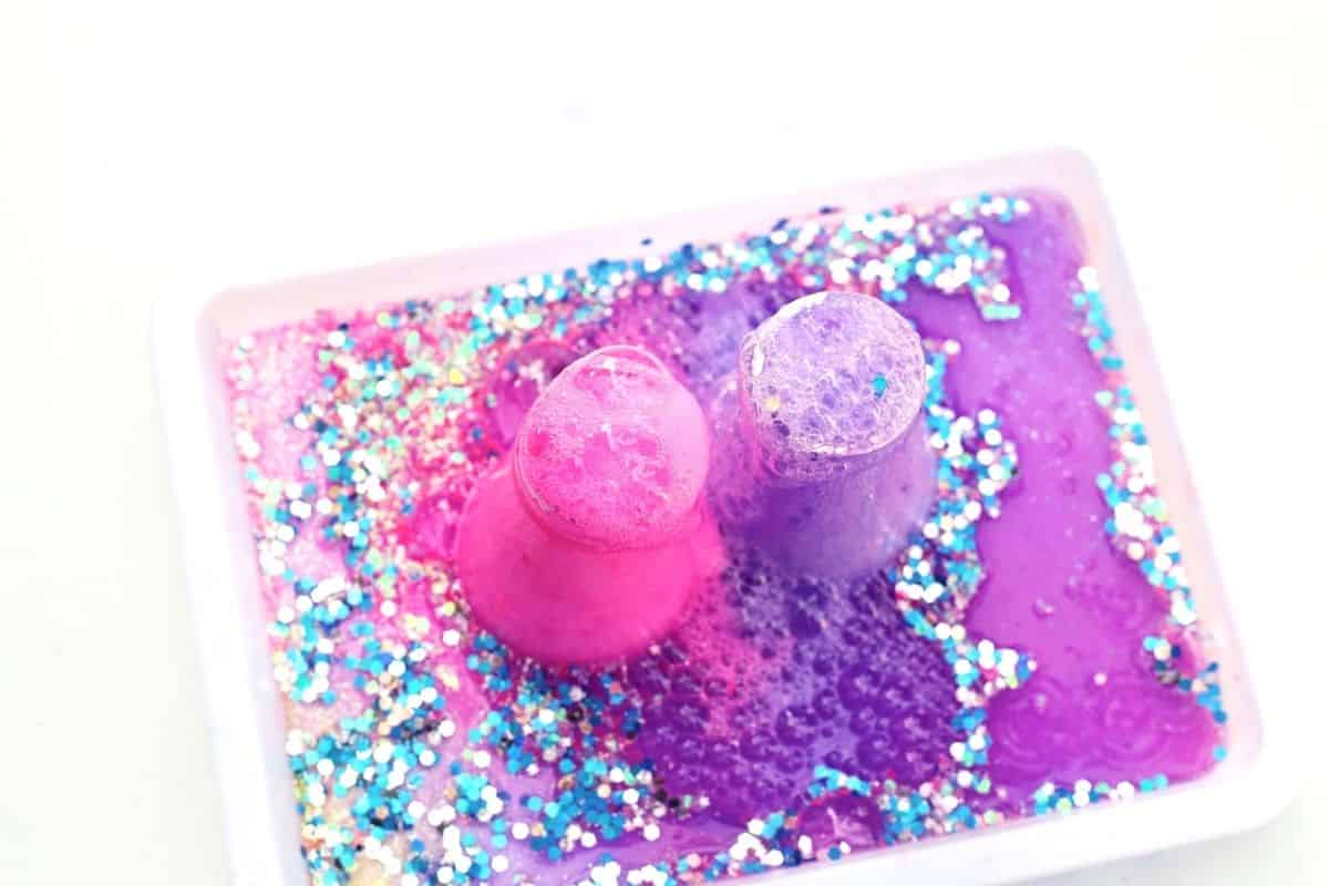 Two small cups overflow with bright pink and purple foam as the fizzy reaction bubbles up inside a white tray filled with blue and silver glitter. The colorful mixture spreads and sparkles, capturing the exciting chemical reaction in this Easter fizzy surprise STEM activity.