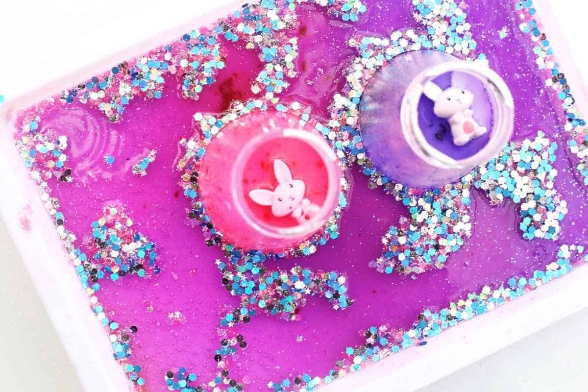 Overhead view of a white tray filled with bright pink and purple fizzy liquid covered in chunky blue and silver glitter, with two small cups holding bunny figurines in the center. The colorful swirling mixture and sparkling texture highlight the final bubbling result of the Easter fizzy surprise STEM activity.