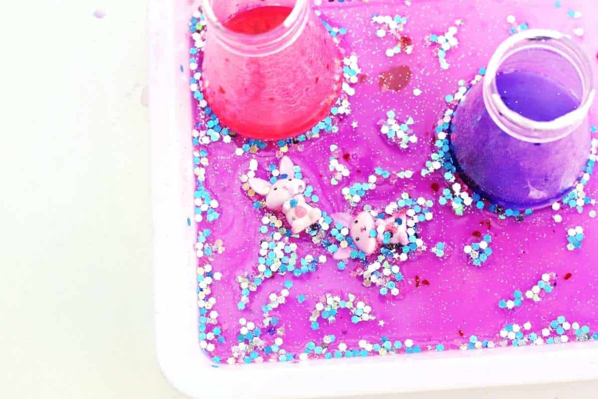 Close up of two small bunny figurines lying in a swirling pink and purple fizzy mixture inside a white tray, surrounded by chunky blue and silver glitter. Two cups filled with bright pink and purple liquid sit nearby, showing the colorful bubbling reaction of the Easter fizzy surprise STEM activity in action.