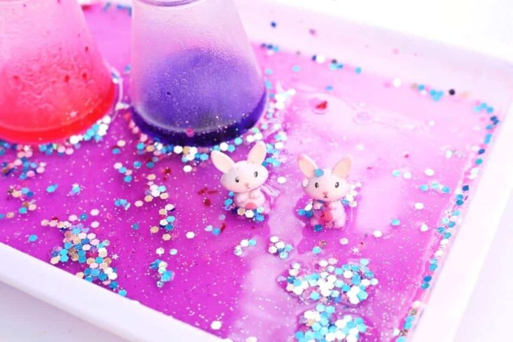 Close up of two small bunny figurines sitting in a pink and purple fizzy mixture filled with blue and silver glitter inside a white tray. Colorful foam rises from two small flasks in the background, showing the bubbling reaction of an Easter fizzy surprise STEM activity in action.