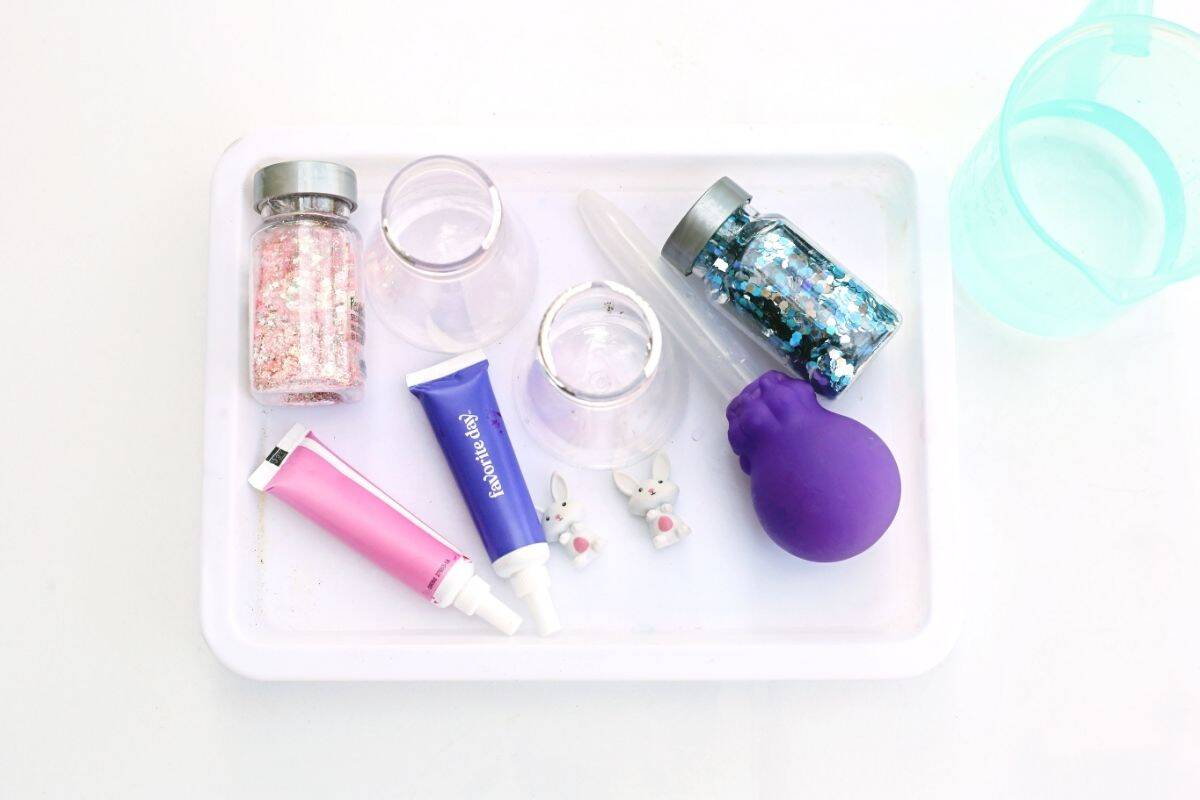 White tray arranged with supplies for an Easter fizzy surprise STEM activity including pink and purple paint tubes, two small bunny figurines, clear plastic cups, pipettes, glitter jars in pink and blue, and a purple balloon. The materials are neatly organized to show everything needed for a colorful hands on Easter science experiment for kids.