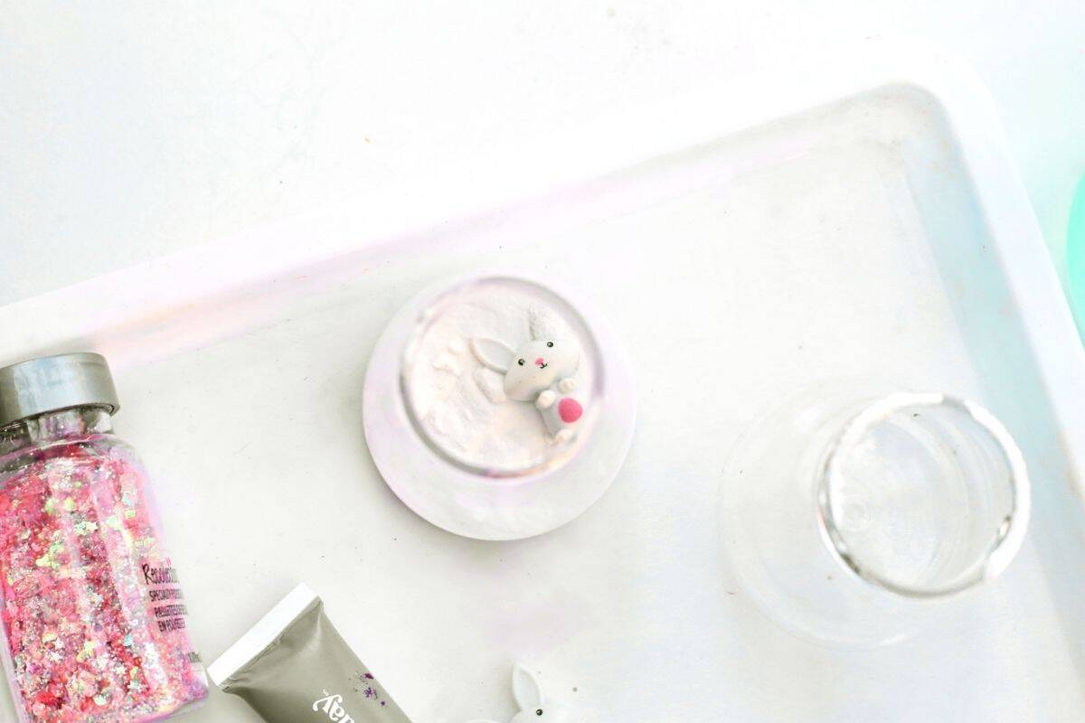 Overhead view of a white tray with a small bunny figurine resting inside a cup filled with white powder, next to an empty clear cup and a jar of pink glitter. The setup shows part of an Easter fizzy surprise STEM activity, highlighting the materials prepared before adding color and creating the bubbling science reaction.
