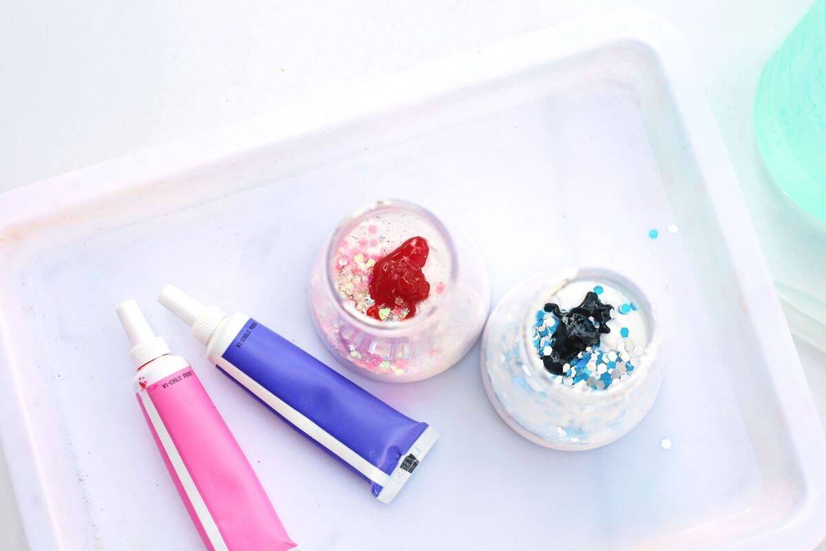 Overhead view of two small cups filled with white powder and glitter on a white tray, with red paint added to one cup and dark purple paint added to the other. Pink and purple paint tubes sit beside the cups, showing the step of adding color in an Easter fizzy surprise STEM activity before the bubbling reaction begins.