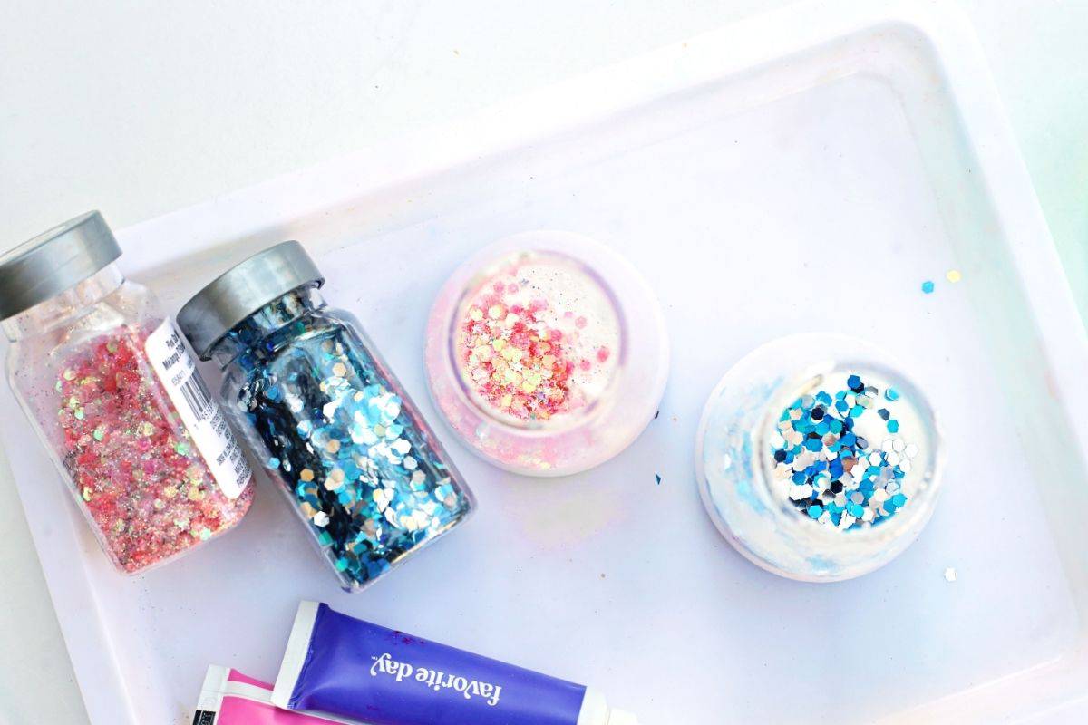 Overhead view of two small cups filled with white powder and topped with pink and blue chunky glitter, placed on a white tray beside jars of pink and blue glitter and tubes of pink and purple paint. The colorful materials are arranged for an Easter fizzy surprise STEM activity, showing the glitter being added before the bubbling science reaction begins.