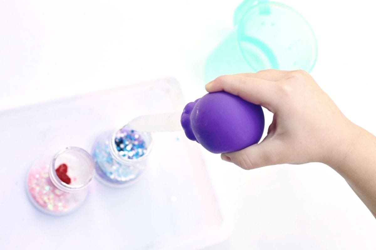 Hand squeezing a purple bulb dropper to add liquid into two small cups filled with white powder and glitter on a white tray. The step shows liquid being added to create the bubbling reaction in an Easter fizzy surprise STEM activity, with pink and blue glitter visible inside the cups.