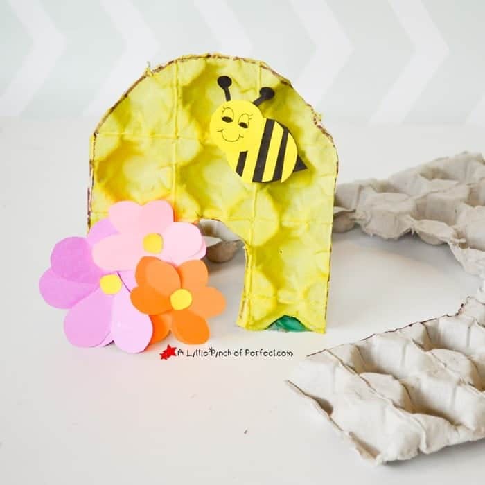 Yellow painted egg carton arch craft decorated like a beehive with a smiling paper bee attached near the top. Pink and orange paper flowers sit at the base on a white surface
