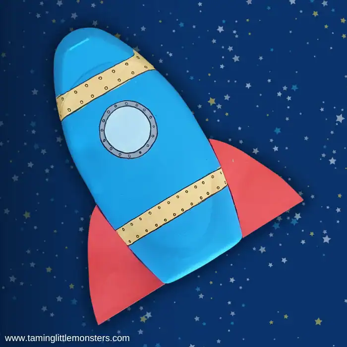 Blue rocket craft made from a painted cardboard tube with red paper fins and a round window, set against a dark blue background filled with small stars.