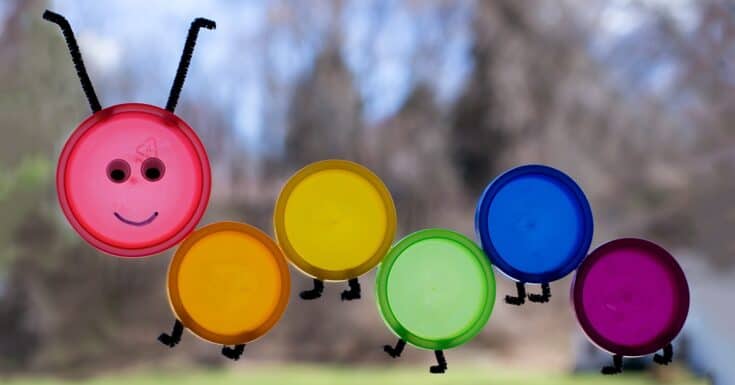 Colorful plastic lid caterpillar craft made from pink, orange, yellow, green, blue, and purple lids arranged in a line outdoors. The pink lid forms the smiling face with black pipe cleaner antennae, and the other lids create the body with small black pipe cleaner legs.