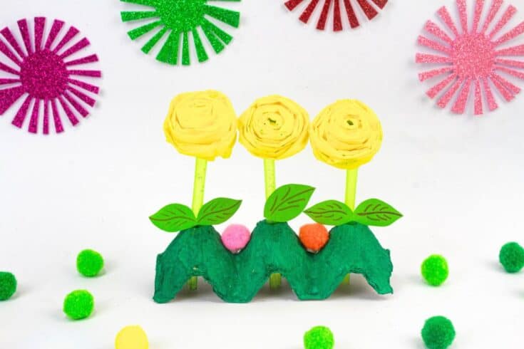 Three yellow paper flowers with rolled spiral tops stand in a green painted egg carton holder on a white background. Glitter foam flower shapes and small pom poms around them make the craft look like a cheerful spring flower display.