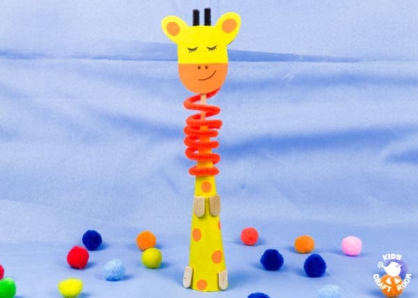 A giraffe themed kids craft stands upright with a yellow paper head and orange muzzle on top of a tall cone body decorated with orange spots. A bright red coil wraps around the neck like a spring and colorful pom poms are scattered on a light blue background