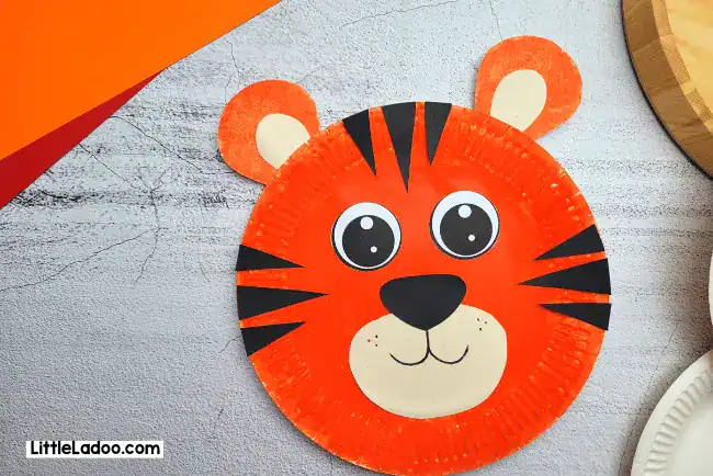 A bright orange paper plate tiger face craft with big cartoon eyes black stripes and a cream muzzle sits on a gray textured background.