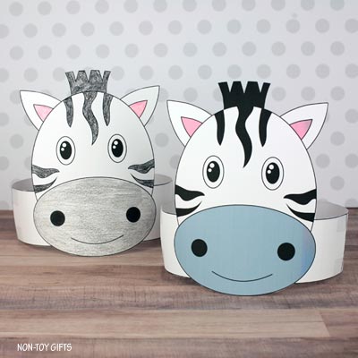 Two printable paper zebra crafts sit on a wooden surface with rounded white bodies black stripes and pink inner ears. One zebra has a gray muzzle and the other has a blue muzzle and both have smiling faces with wide cartoon eyes.