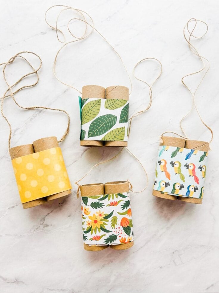 Four homemade binocular crafts made from cardboard tubes are wrapped in patterned paper and tied with string on a white marble surface. The designs include yellow polka dots, green leaves, tropical flowers, and colorful birds, showing simple recycled craft ideas for kids.