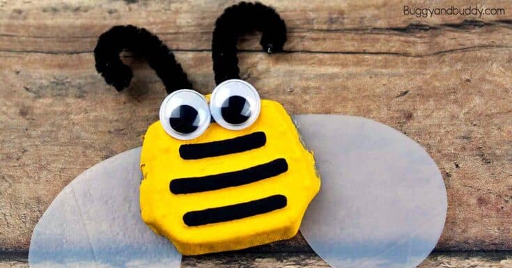 Yellow bee craft with black pipe cleaner antennae, large googly eyes, and three black stripes placed on a rustic wooden surface. The bee has translucent wings spread out on both sides and looks like a simple kid made art project.