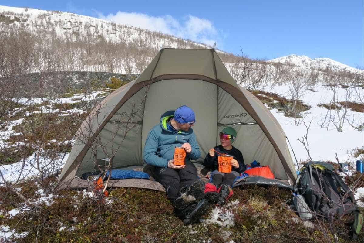An adult and child sit at the opening of a tent in a snowy mountain landscape, eating from bright orange meal pouches and looking at each other. Backpacks and camping gear rest beside them, with patches of snow, low brush, and white peaks stretching behind the campsite.