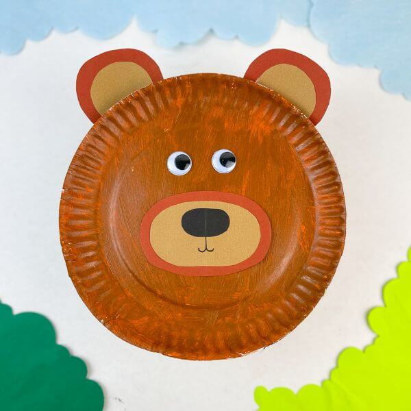 A painted paper plate bear face craft with googly eyes and a tan snout is centered on a white background. Blue cloud shapes and green paper bushes frame the bear like a simple forest scene.