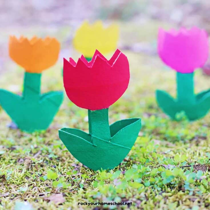 Bright paper tulip crafts in red, orange, yellow, and pink stand upright on mossy ground with a soft blurred background. The red tulip is centered in front with folded green leaves and stem, while the others sit behind it like a simple spring kids craft display.