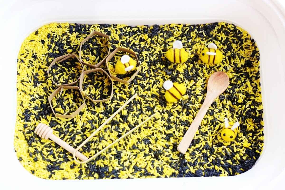 Overhead view of a bee sensory bin filled with yellow and black dyed rice with cardboard hexagon honeycomb shapes, felt bees, a wooden spoon, honey dipper, and bamboo tongs arranged for sensory play and scooping activities.