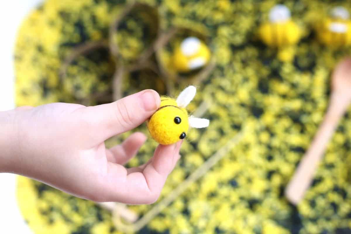 A child’s hand holds a small felt bee with white wings above a bee sensory bin filled with yellow and black dyed rice, with honeycomb shapes and wooden tools blurred in the background to show hands on play.