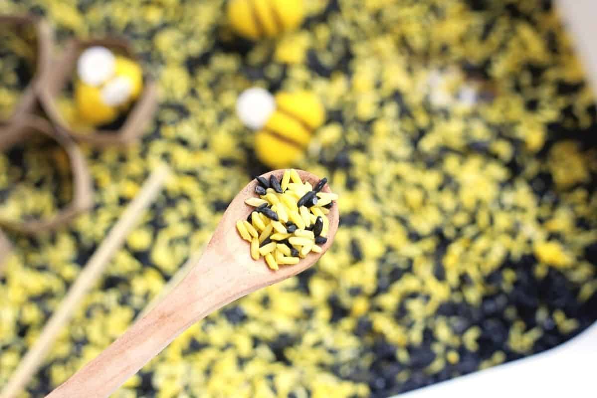 A wooden spoon holds a scoop of yellow and black dyed rice above a bee sensory bin with felt bees and cardboard honeycomb shapes blurred in the background to highlight scooping and pouring play.