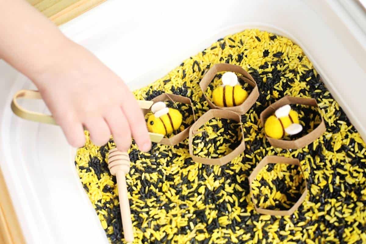 A child uses wooden tongs and a honey dipper to move felt bees into cardboard honeycomb shapes in a bee sensory bin filled with yellow and black dyed rice.