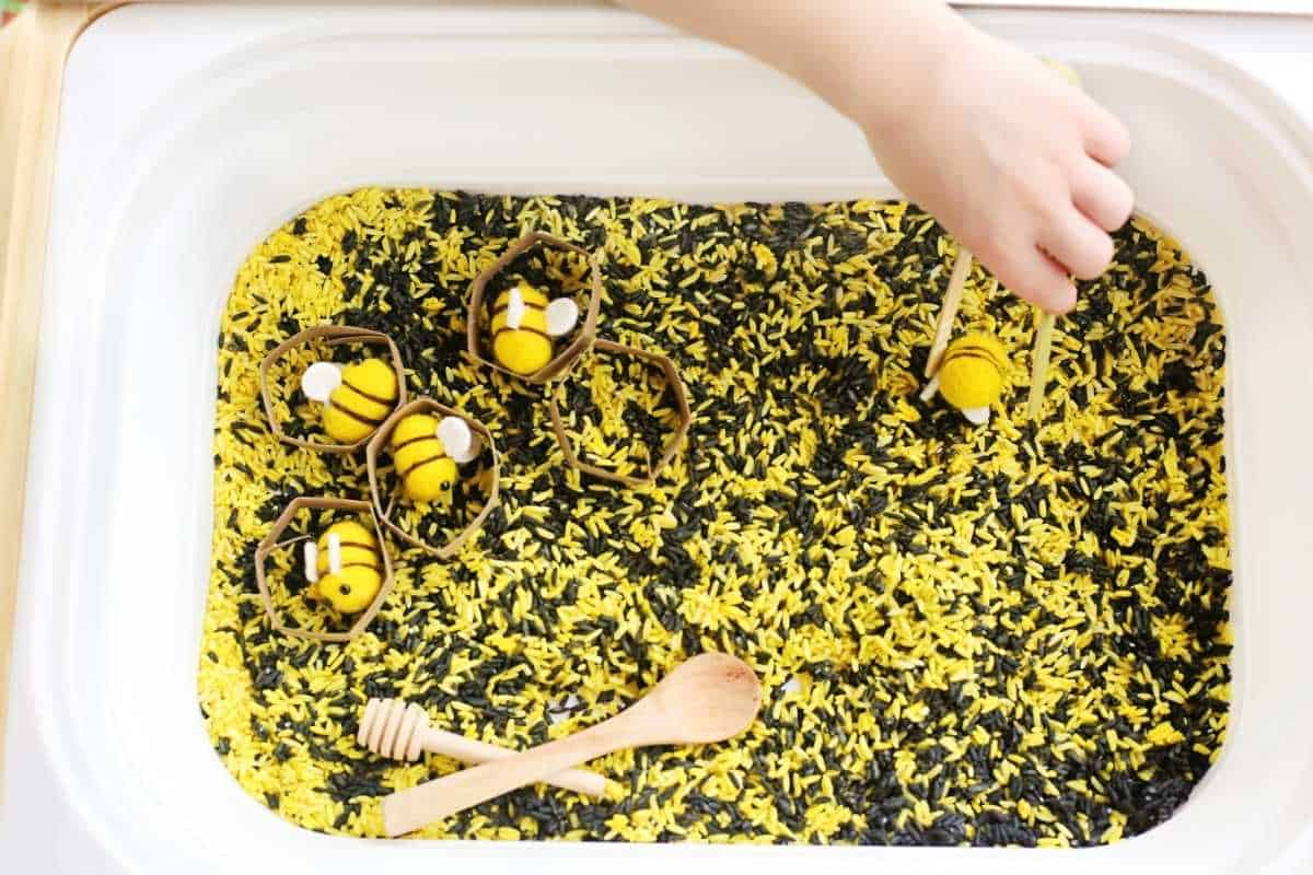 A child uses wooden tongs to pick up a felt bee and place it into cardboard honeycomb shapes in a bee sensory bin filled with yellow and black dyed rice, with a wooden spoon and honey dipper nearby.