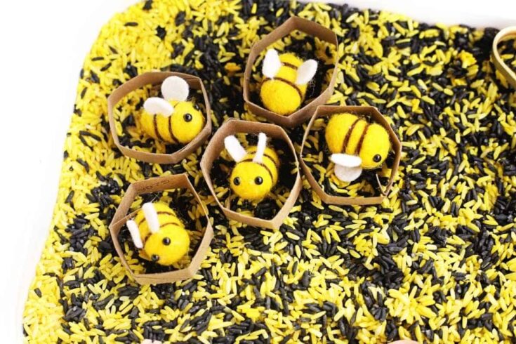 Felt bees sit inside small cardboard hexagon honeycomb shapes on top of yellow and black dyed rice in a bee sensory bin, showing a simple sorting and imaginative play setup.