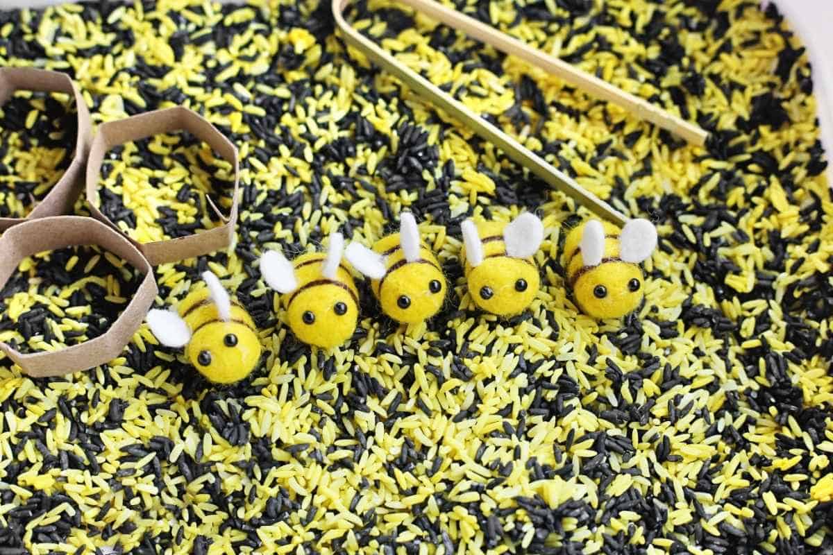 Five small felt bees are lined up on yellow and black dyed rice inside a bee sensory bin with cardboard honeycomb shapes and bamboo tongs nearby for sorting and imaginative play.
