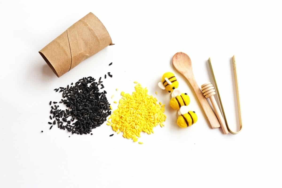 Materials for a bee sensory bin including black and yellow dyed rice, small felt bees, a cardboard tube, wooden spoon, honey dipper, and bamboo tongs arranged on a white surface for a hands on sensory play setup.