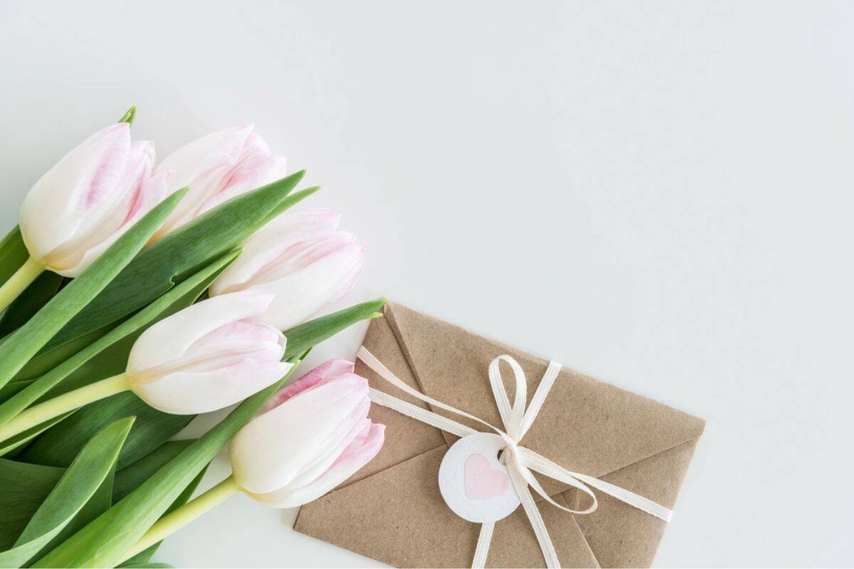 Soft pink tulips with green leaves rest beside a brown envelope tied with white string and a small heart tag on a clean white background.