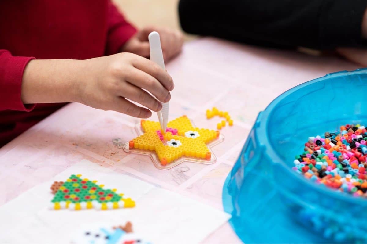 Child using tweezers to place colorful beads onto a star shaped pegboard craft while sitting at a table with a container of beads nearby. A partially finished bead design and printed pattern sheet are visible beside the child’s hands.