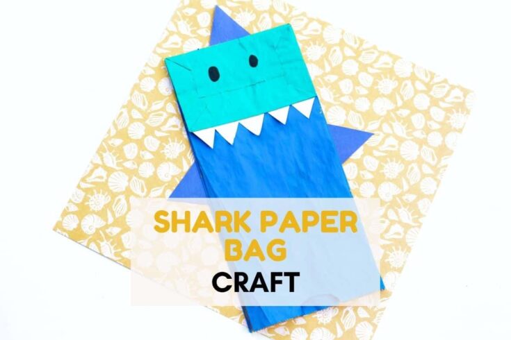 Blue shark paper bag craft with a teal shark head, white triangle teeth, and blue fins placed on patterned paper with the text “SHARK PAPER BAG CRAFT” across the center.