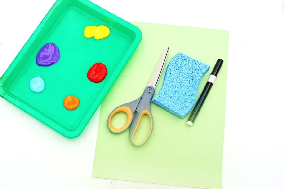 Sponge painted caterpillar supplies-2 Supplies for a sponge painted caterpillar craft including colorful paint on a green tray, scissors, a blue sponge, black marker, and light green paper laid out on a white table. Purple, red, blue, orange, and yellow paint are arranged for making the sponge painted caterpillar art activity.